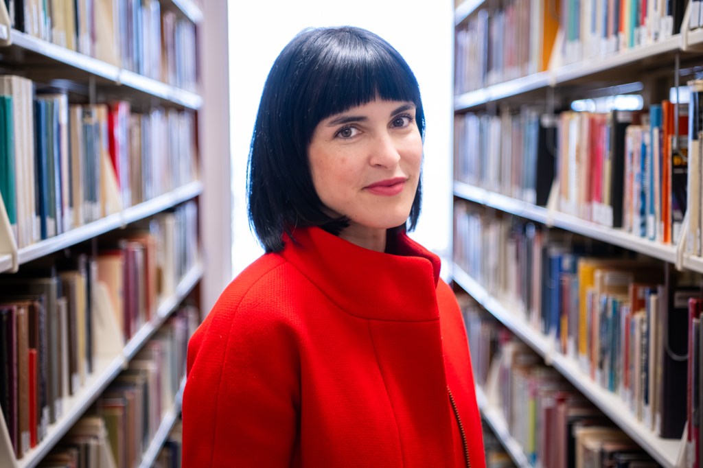 Headshot of Keegan Finberg, woman with dark hair standing in library in red coat.